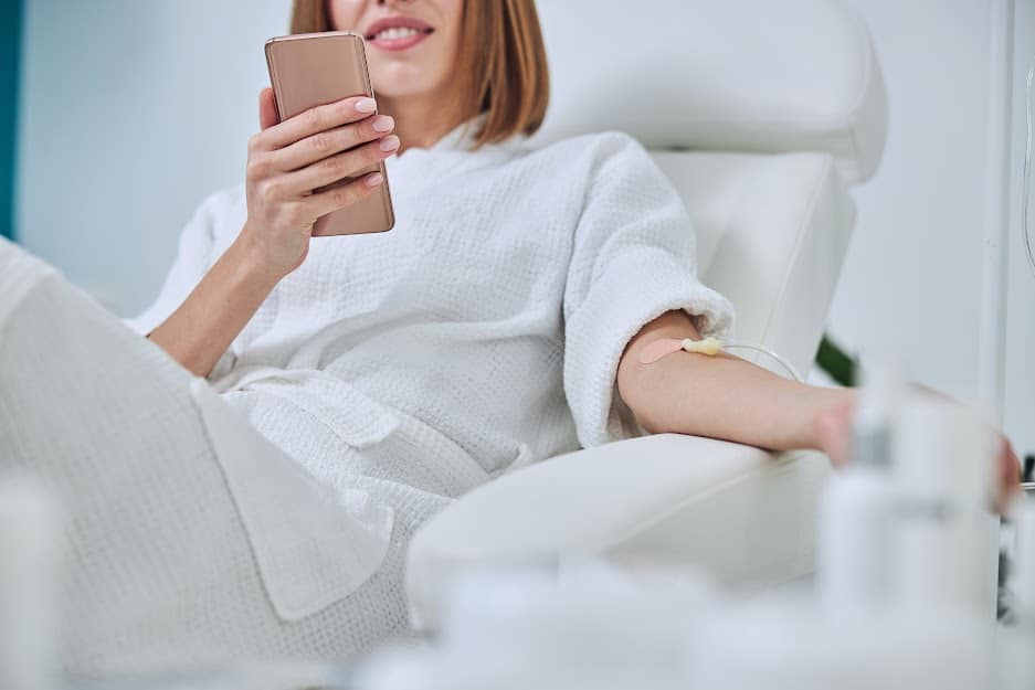 Woman in hospital talking at mobile phone while needle is in arm, Vitamin Therapy Iv Drip Infusion In Women Blood | Instinct Holistic Medical Spa IN Bozeman, MT