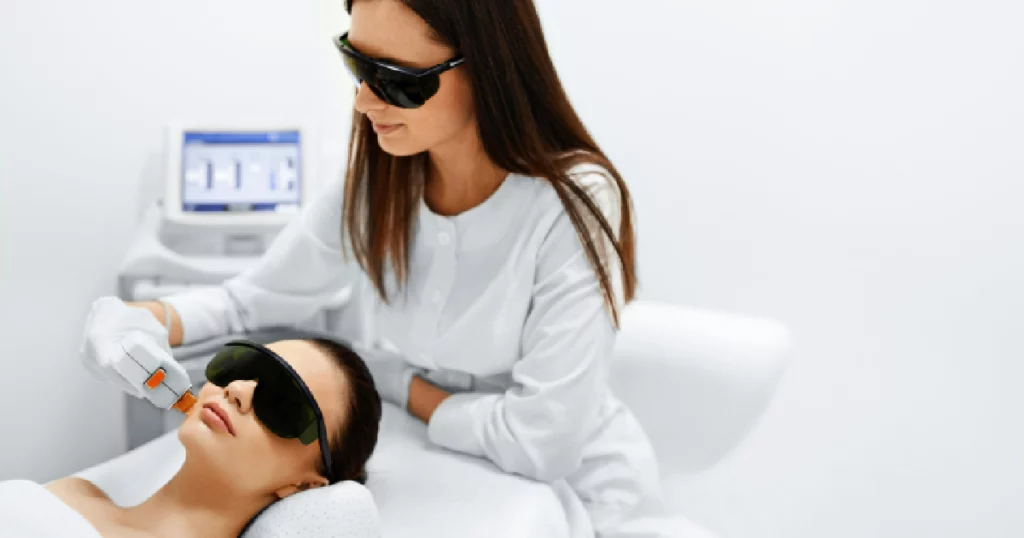 A skincare specialist performing a photofacial treatment on a female client using IPL (Intense Pulsed Light) technology. Both are wearing protective eyewear in a clinical setting in BOZEMAN, MT